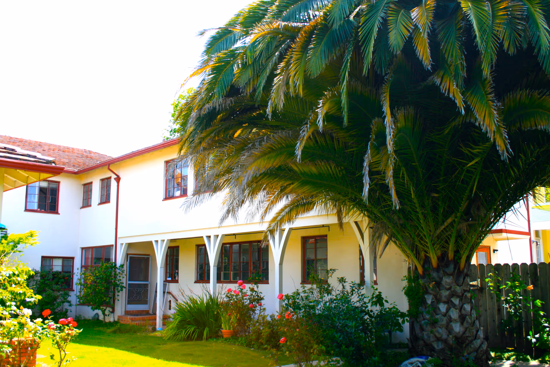 Two-story white building with a covered porch, a large palm tree and a flowering front garden.