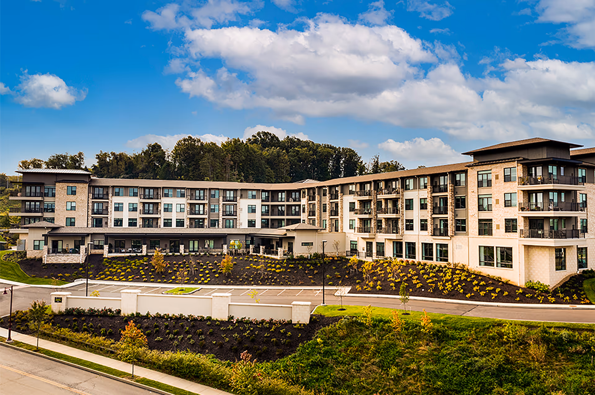 Exterior view of a large, modern senior living facility building with multiple floors, balconies, and large windows. The building is surrounded by landscaped grounds with small trees and shrubs, and a parking area is visible in front. The sky is partly cloudy with blue patches.