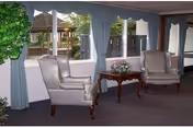 A lounge with two white armchairs, a wooden table with flowers, and blue curtains by windows.