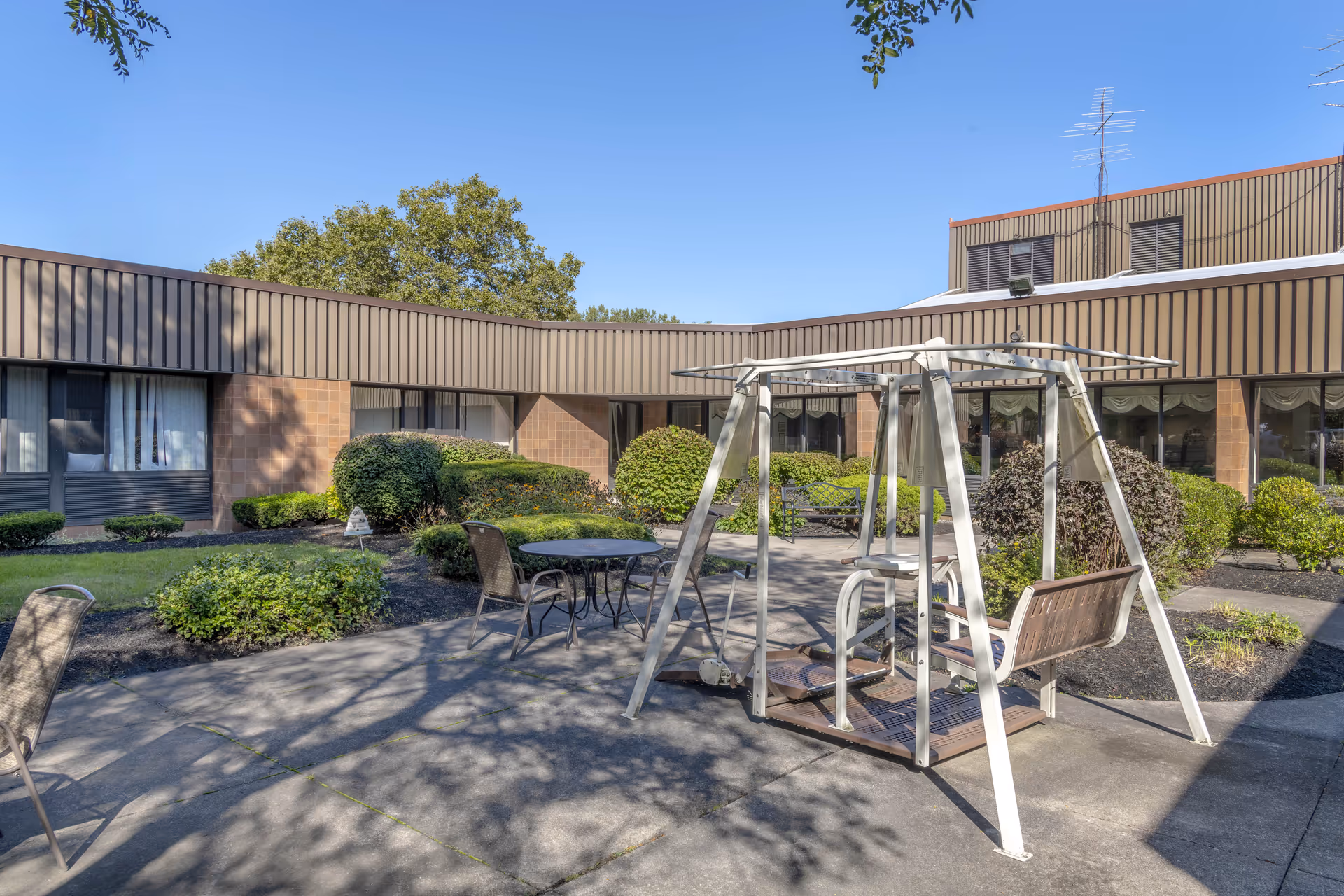 Outdoor courtyard area at a senior living facility with a metal swing bench, several chairs, and a round table surrounded by bushes and greenery. The building with windows and brown paneling encloses the courtyard under a clear blue sky.