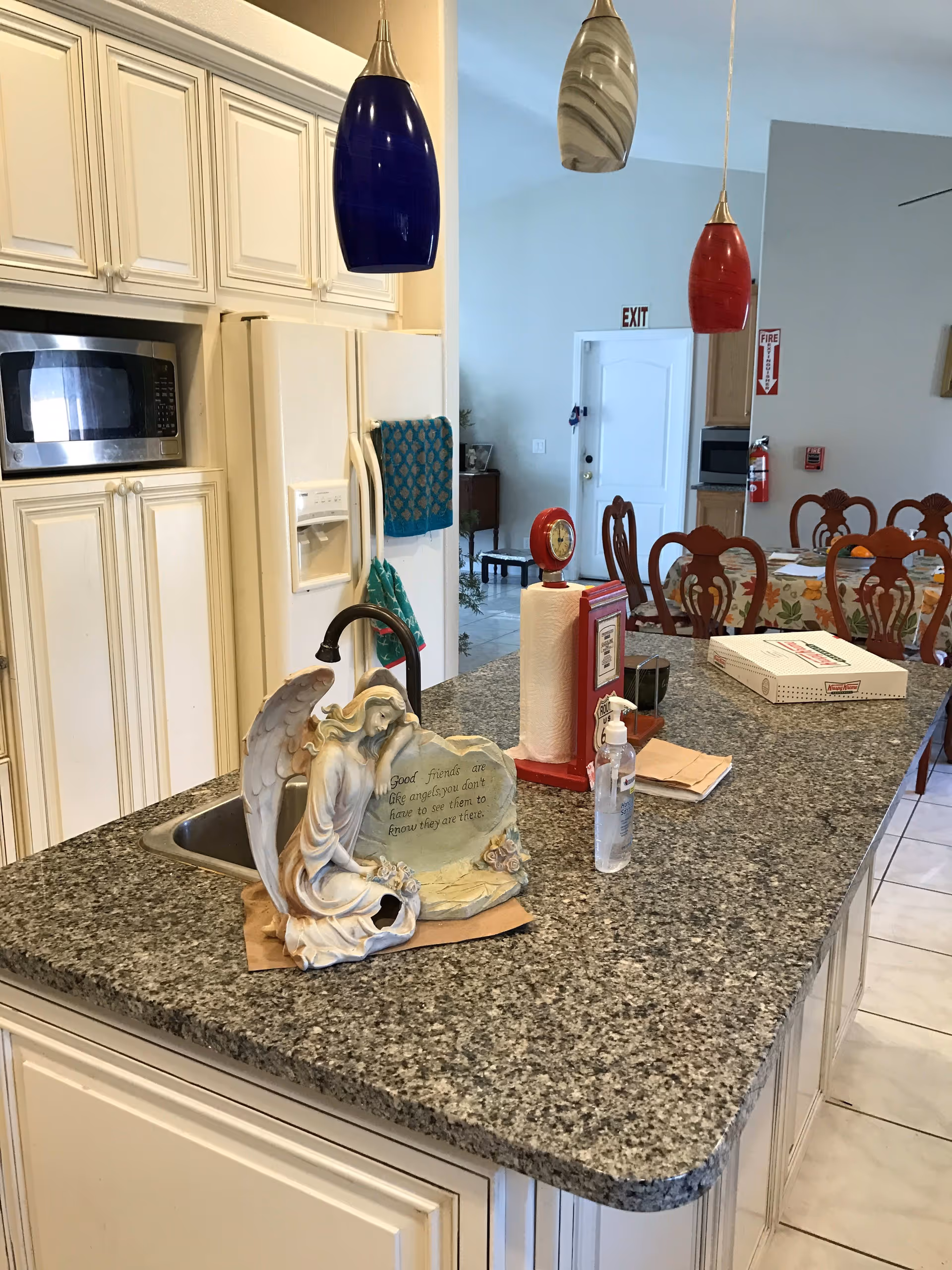 Kitchen island with a granite countertop and sink topped by pendant lights, an angel statue, paper towels and other items, with a refrigerator and dining chairs visible in the background.