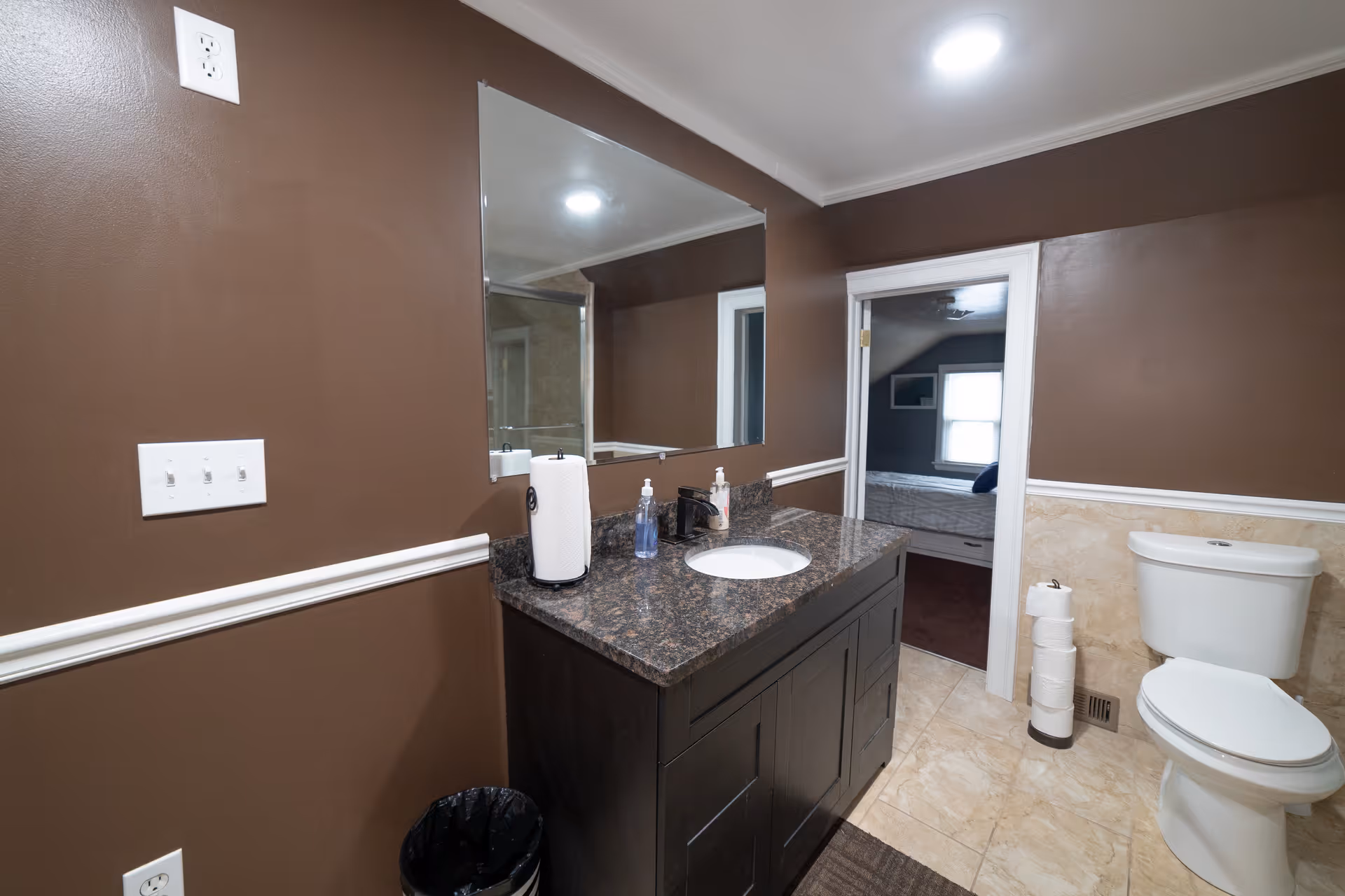 Brown-walled bathroom with granite vanity, mirror, toilet, and doorway to a bedroom.