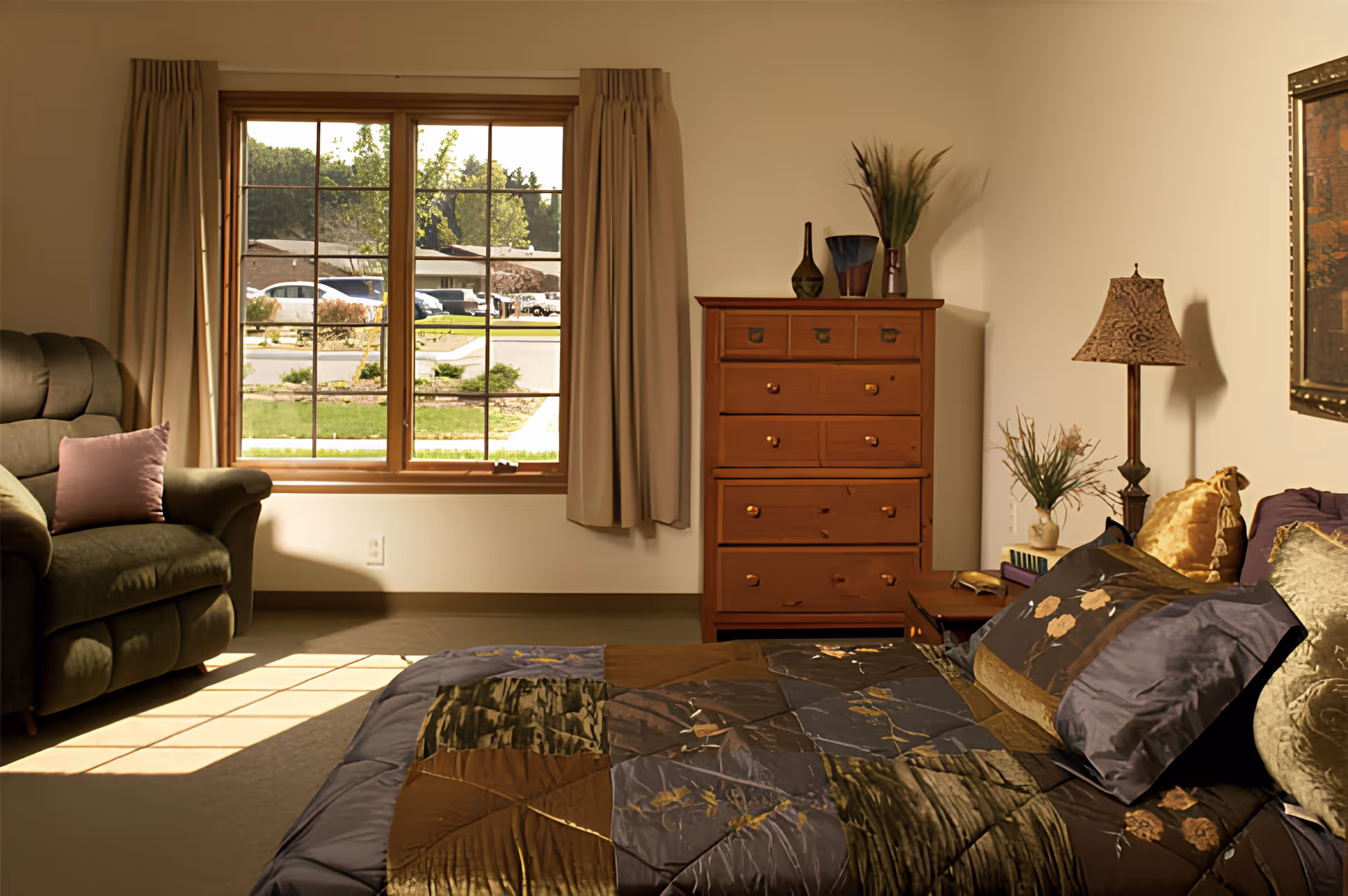 Sunlit bedroom with a bed, wooden dresser, armchair and a large window looking out to the street.