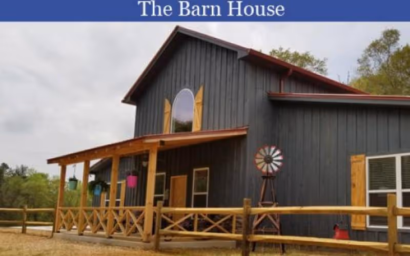 Exterior view of a rustic barn-style house with dark wooden siding and a red roof. The house features a covered porch with wooden railings and hanging flower pots. A small decorative windmill is positioned near the porch. The sky is cloudy and trees are visible in the background.