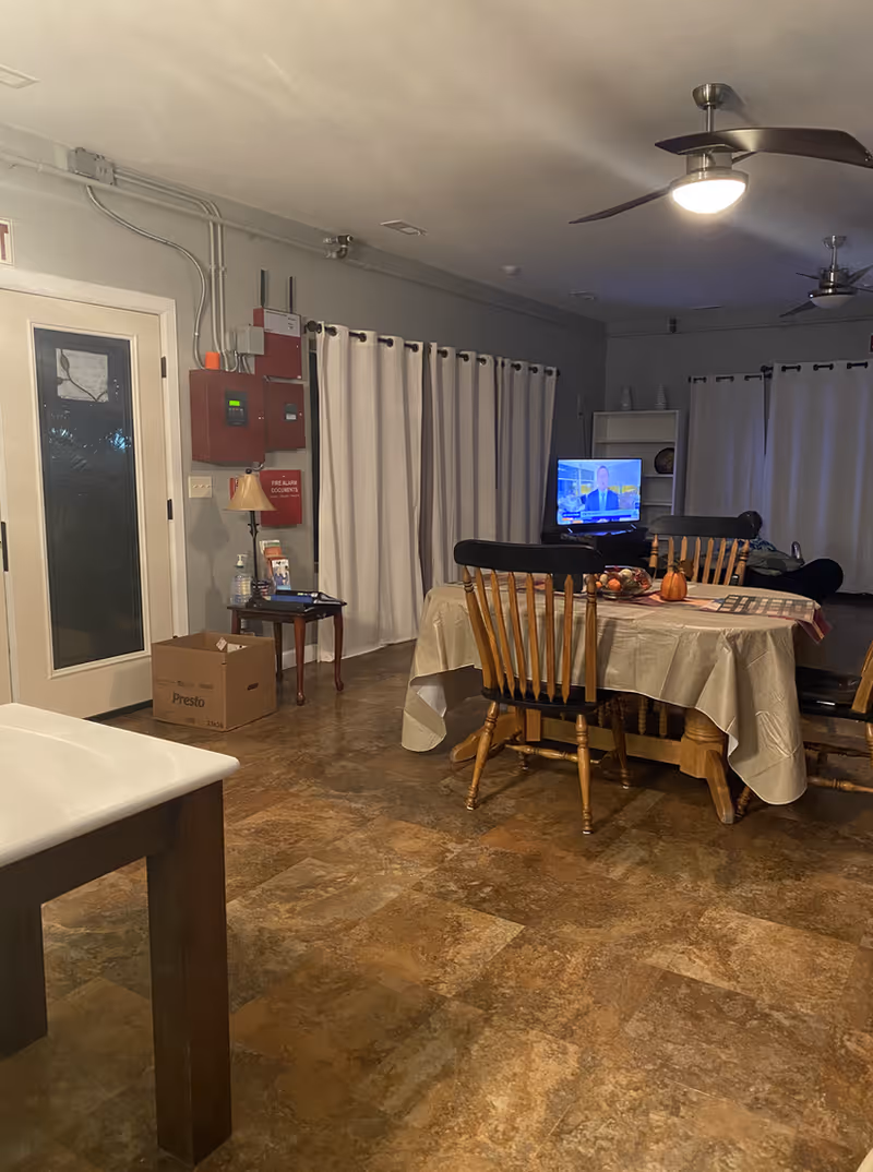 Interior view of a room with a dining table covered with a beige tablecloth and surrounded by wooden chairs. A television is on in the background, placed on a stand near a white bookshelf. The room has beige curtains covering the windows, ceiling fans with lights, and a tiled floor. A cardboard box and a small table with a lamp and some items are near a door on the left side.