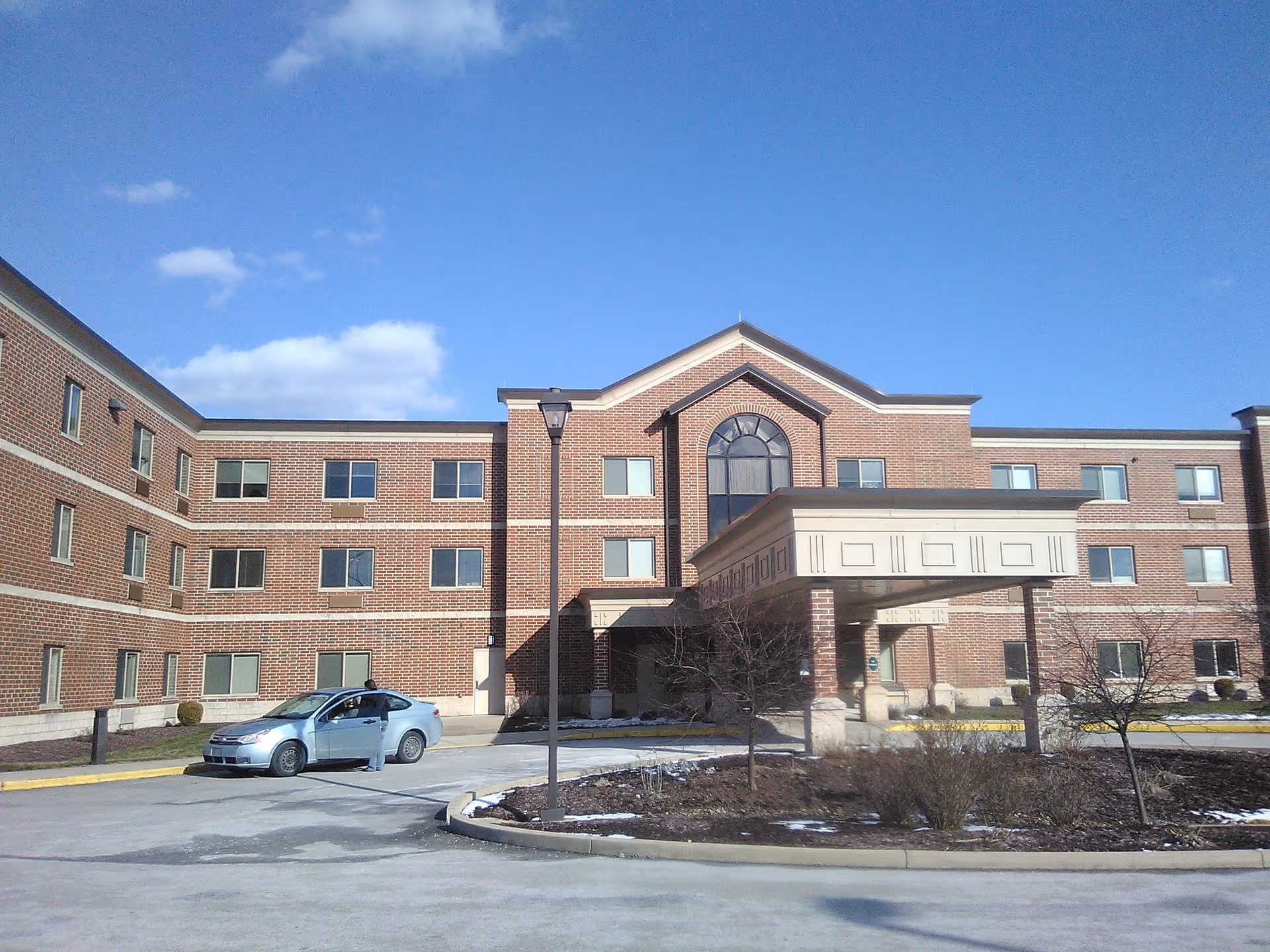 Exterior brick senior living building with covered entrance, driveway, and a parked car.