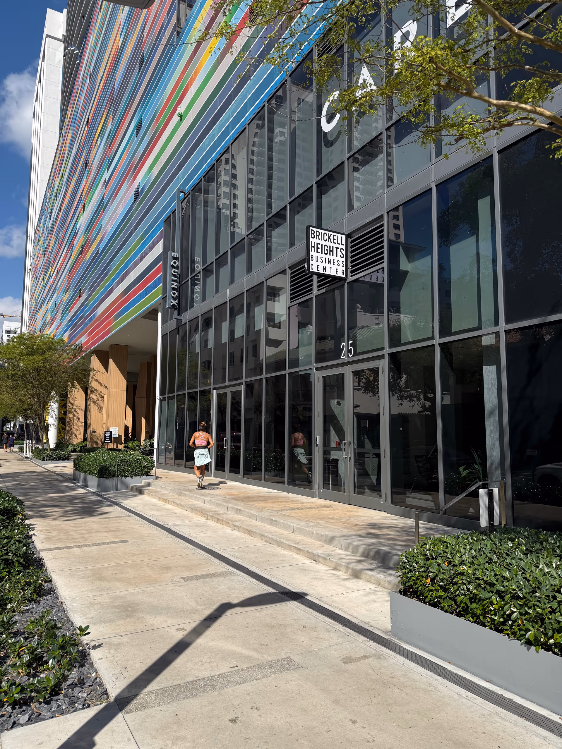 Modern glass-front building with colorful stripes and a sidewalk entry, two pedestrians nearby.