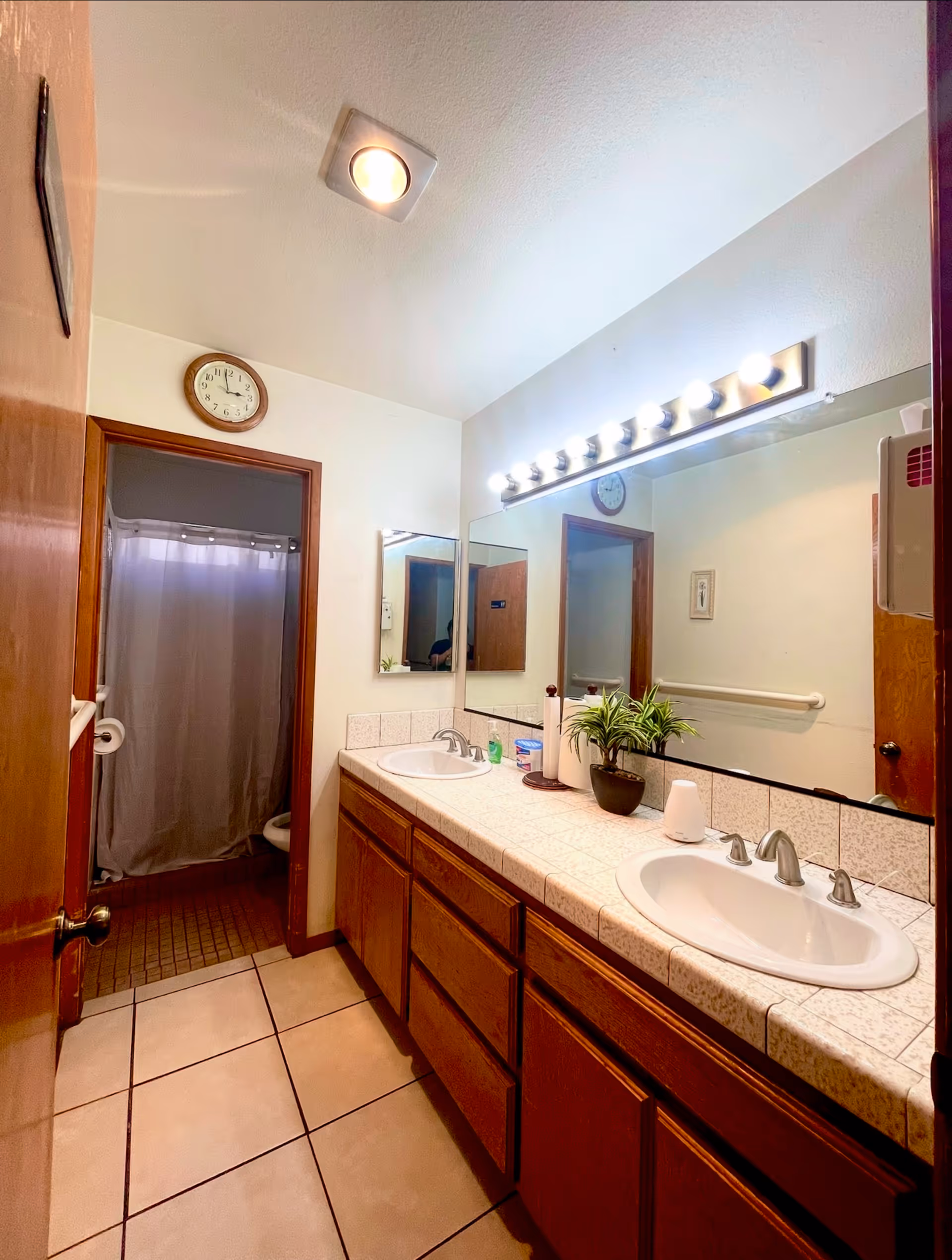 A bathroom with a double sink vanity featuring wooden cabinets and a tiled countertop. Above the sinks is a large mirror with bright vanity lights. A small potted plant and various toiletries are on the counter. In the background, there is a doorway leading to a shower area with a curtain and a toilet partially visible. The floor is tiled, and a clock is mounted on the wall above the doorway.