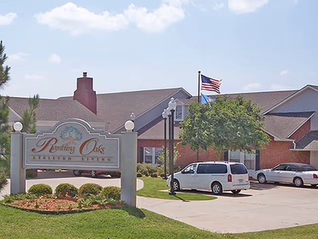 Exterior view of Wickshire Senior Living Oklahoma City entrance with sign, flag, and parked cars.