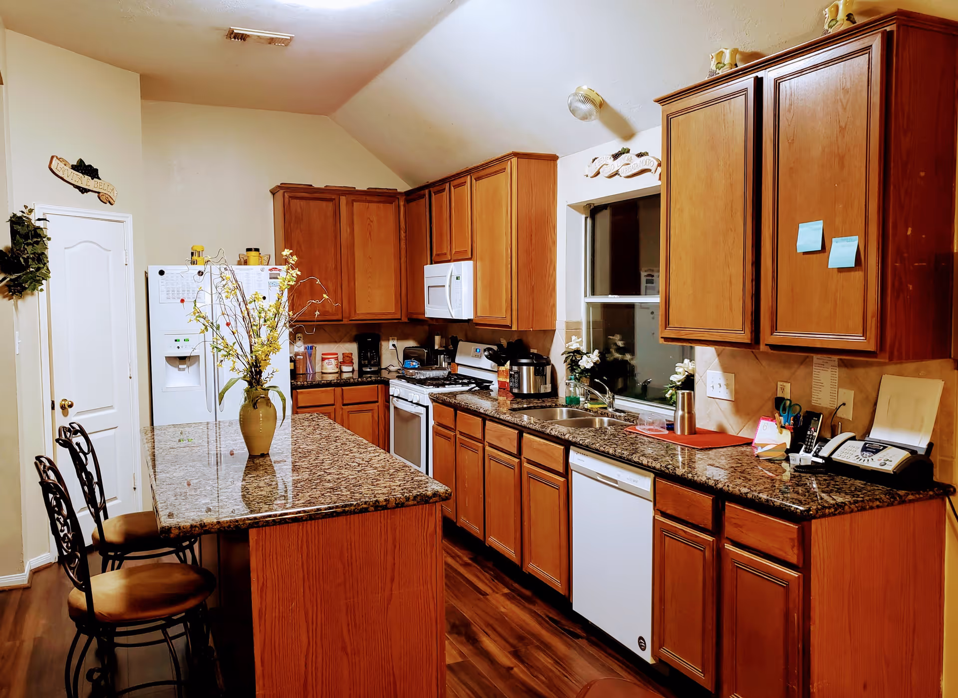 A well-lit kitchen with wooden cabinets, granite countertops, and a kitchen island with two chairs. The kitchen features a white refrigerator, microwave, stove, dishwasher, and a double sink under a window. Various kitchen items and decorations are placed on the countertops and cabinets.