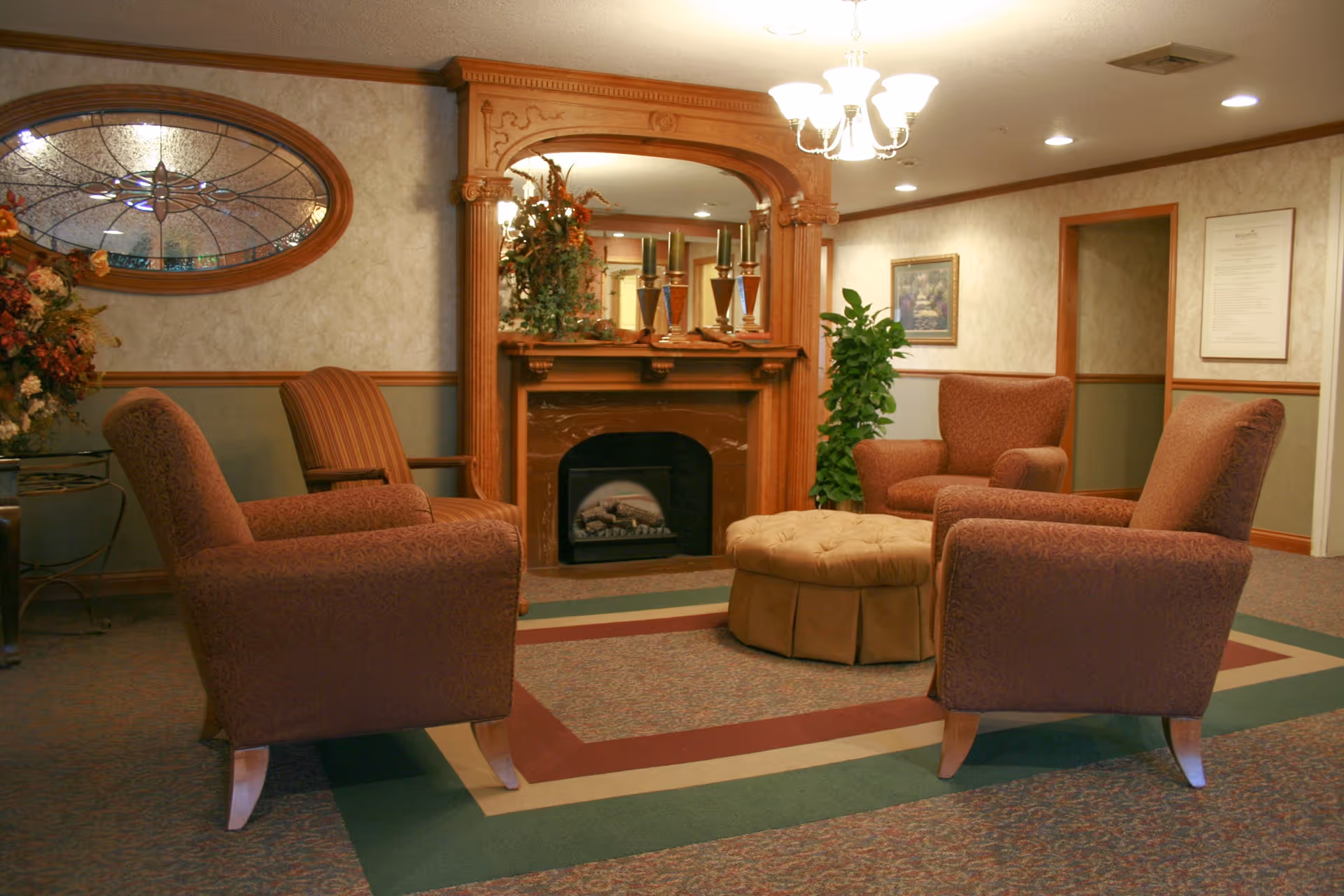 A cozy sitting area with four upholstered armchairs arranged around an ottoman in front of a decorative fireplace and stained-glass window.