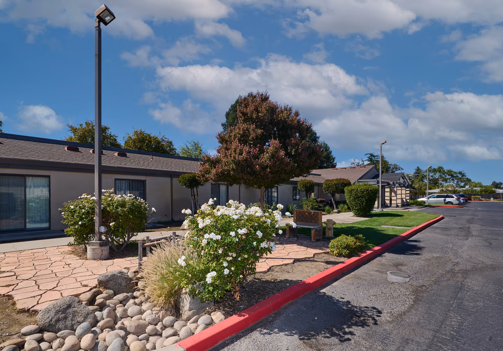 Front courtyard of a single-story residential building with flowering shrubs, a bench, lamp posts and a parking lot under a blue sky.