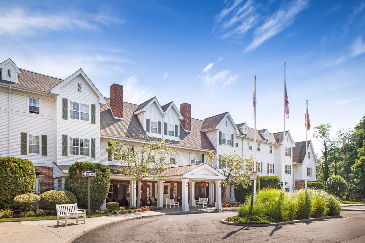 White multi-story senior living building with peaked roofs, flags, and a covered entrance.