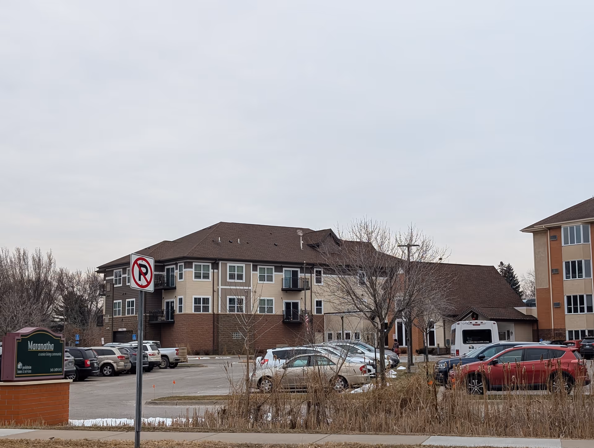 Exterior view of Maranatha Care Center, a senior living community building with a parking lot in front. Several cars are parked, and leafless trees are visible around the area under a cloudy sky.