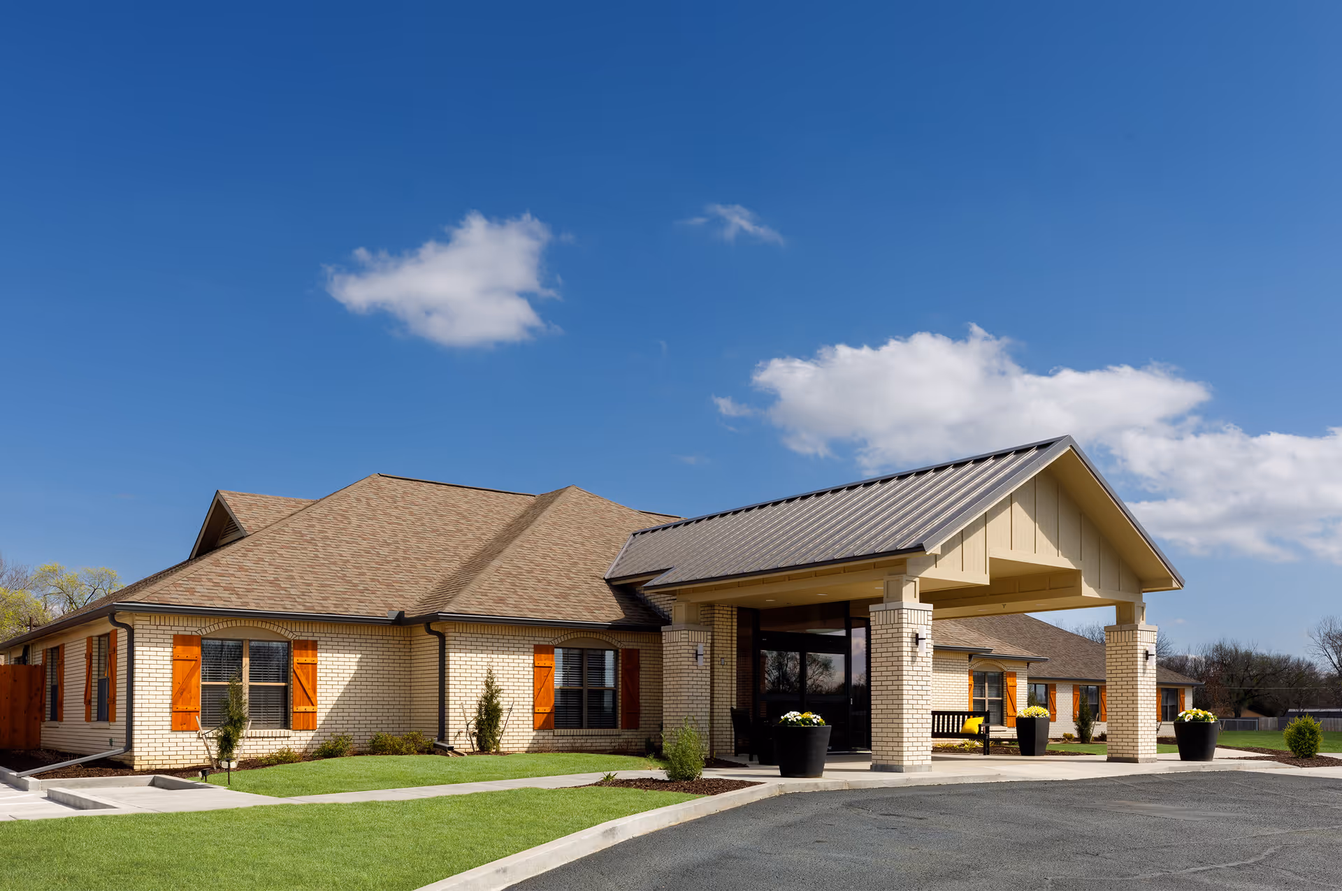 Exterior view of a single-story senior living facility building with beige brick walls, brown shingled roof, and a covered entrance supported by columns. The building has windows with wooden shutters and is surrounded by a well-maintained lawn and paved driveway under a clear blue sky with a few clouds.