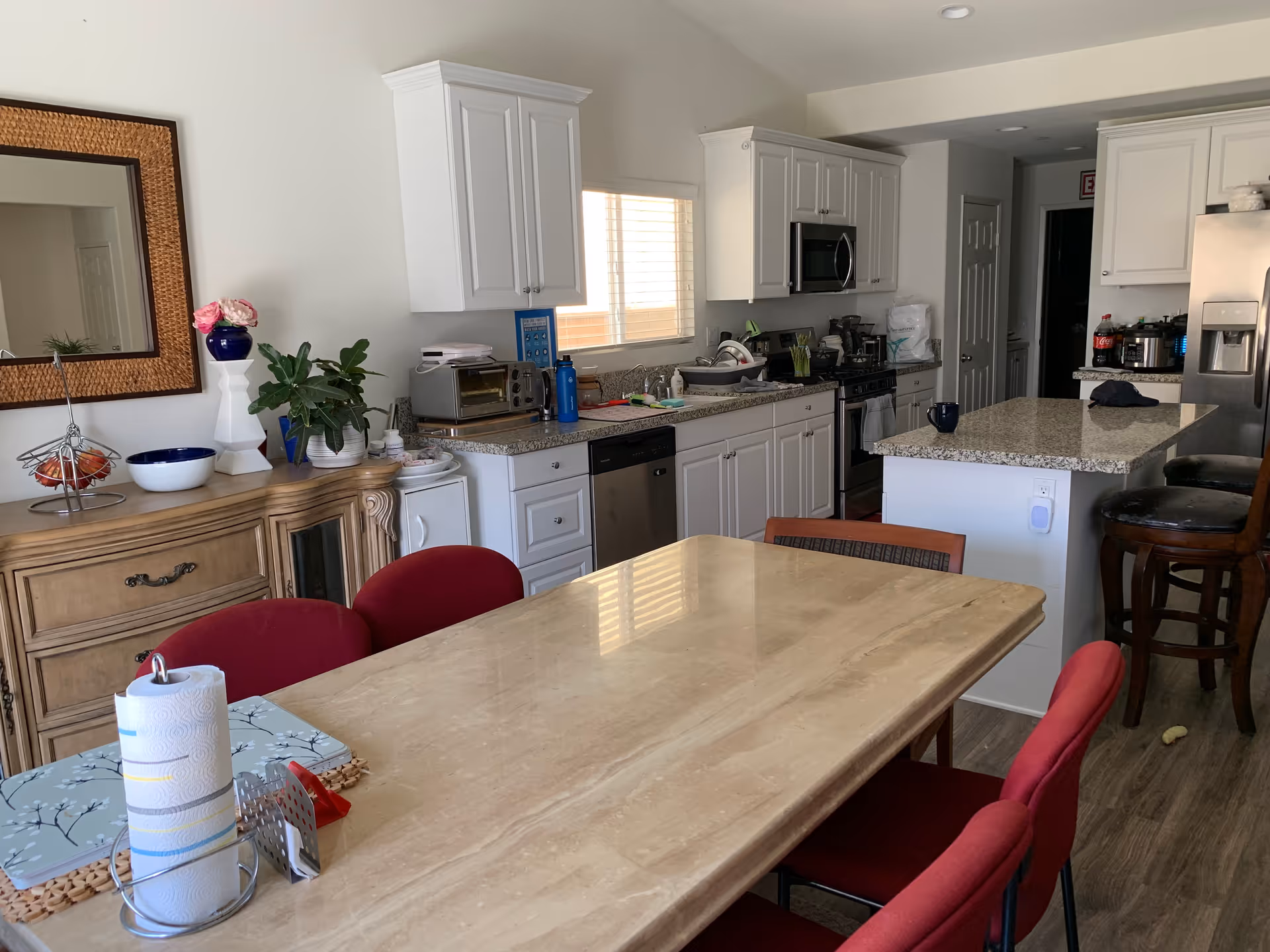 Interior view of a kitchen and dining area with a beige marble dining table surrounded by red chairs. The kitchen features white cabinets, granite countertops, a stainless steel dishwasher, microwave, stove, and refrigerator. There is a wooden sideboard with a mirror, plants, and decorative items on the left side of the image.