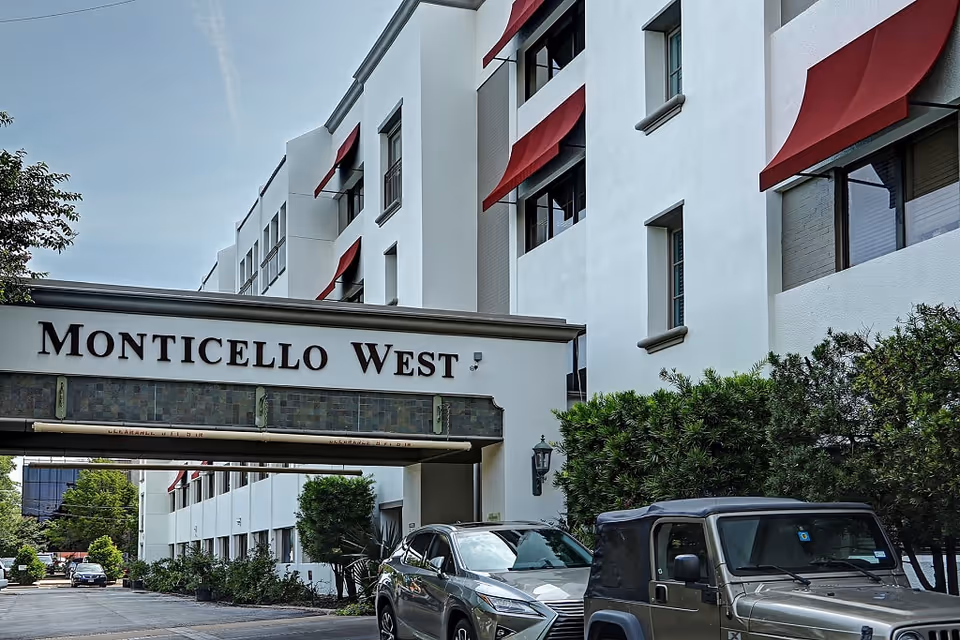 Exterior view of Monticello West building with white walls and red window awnings. There is a covered entrance with the name Monticello West displayed prominently. Several cars are parked in front of the building along a driveway lined with greenery.