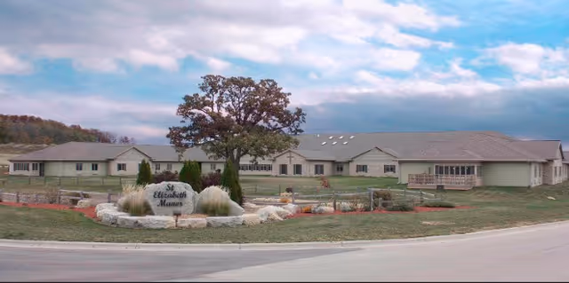 Single-story ranch-style senior living building with a landscaped circular entrance and a sign in front under a partly cloudy sky.