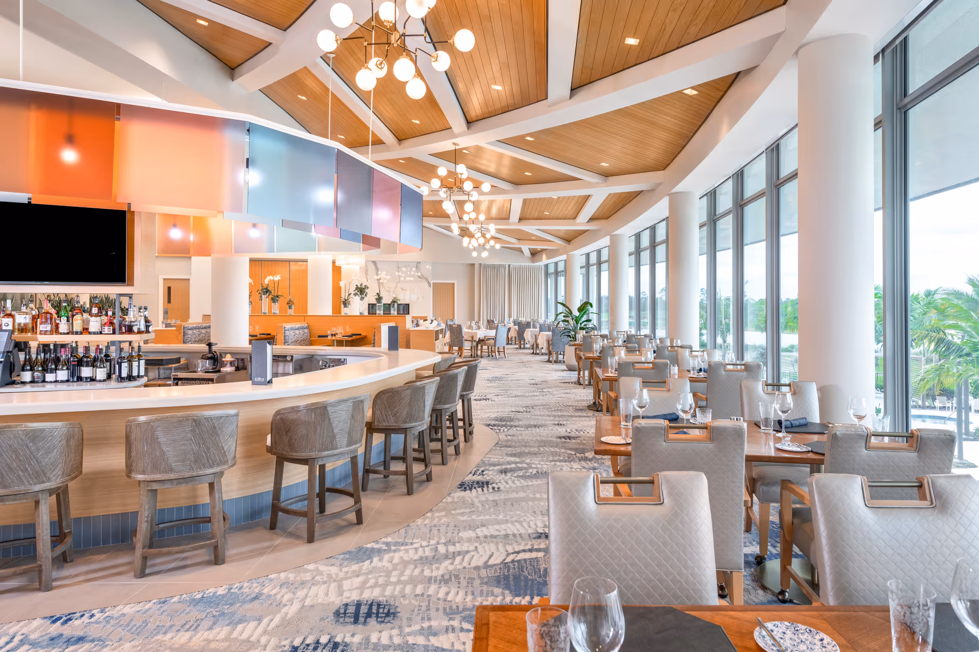 A spacious, modern dining area with large floor-to-ceiling windows allowing natural light to fill the room. The room features a long curved bar with wooden bar stools, colorful hanging panels above the bar, and multiple dining tables with cushioned chairs set with glassware and plates. The ceiling has wooden panels with white beams and contemporary light fixtures.