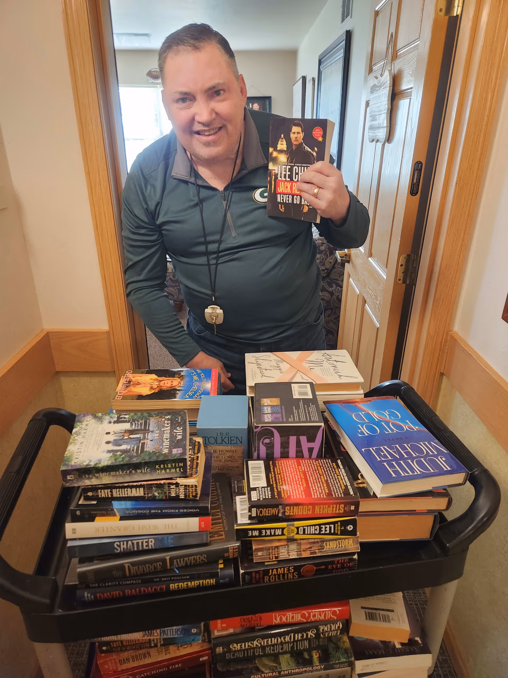 A man wearing a green long-sleeve shirt stands behind a cart filled with a large stack of books in a hallway. He is smiling and holding up a book titled 'Jack Reacher: Never Go Back' by Lee Child. The hallway has light-colored walls and wooden doors.