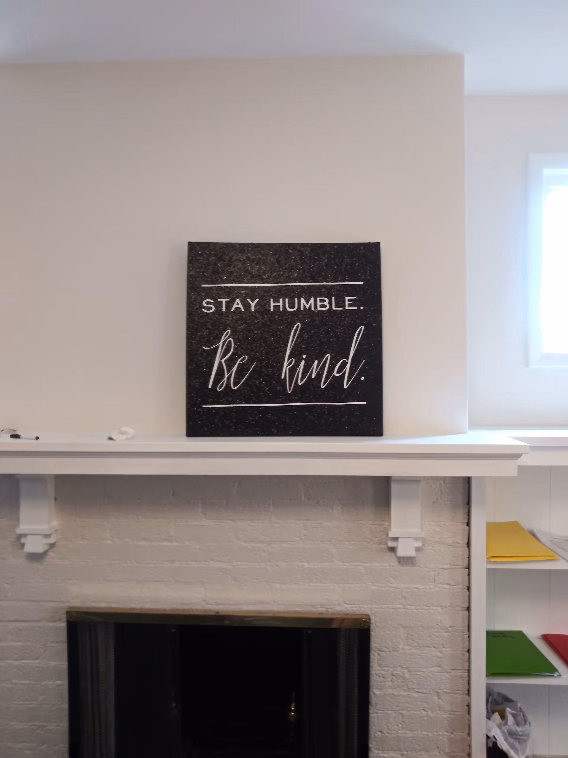 White fireplace mantel with a black sign reading Stay Humble, Be Kind.