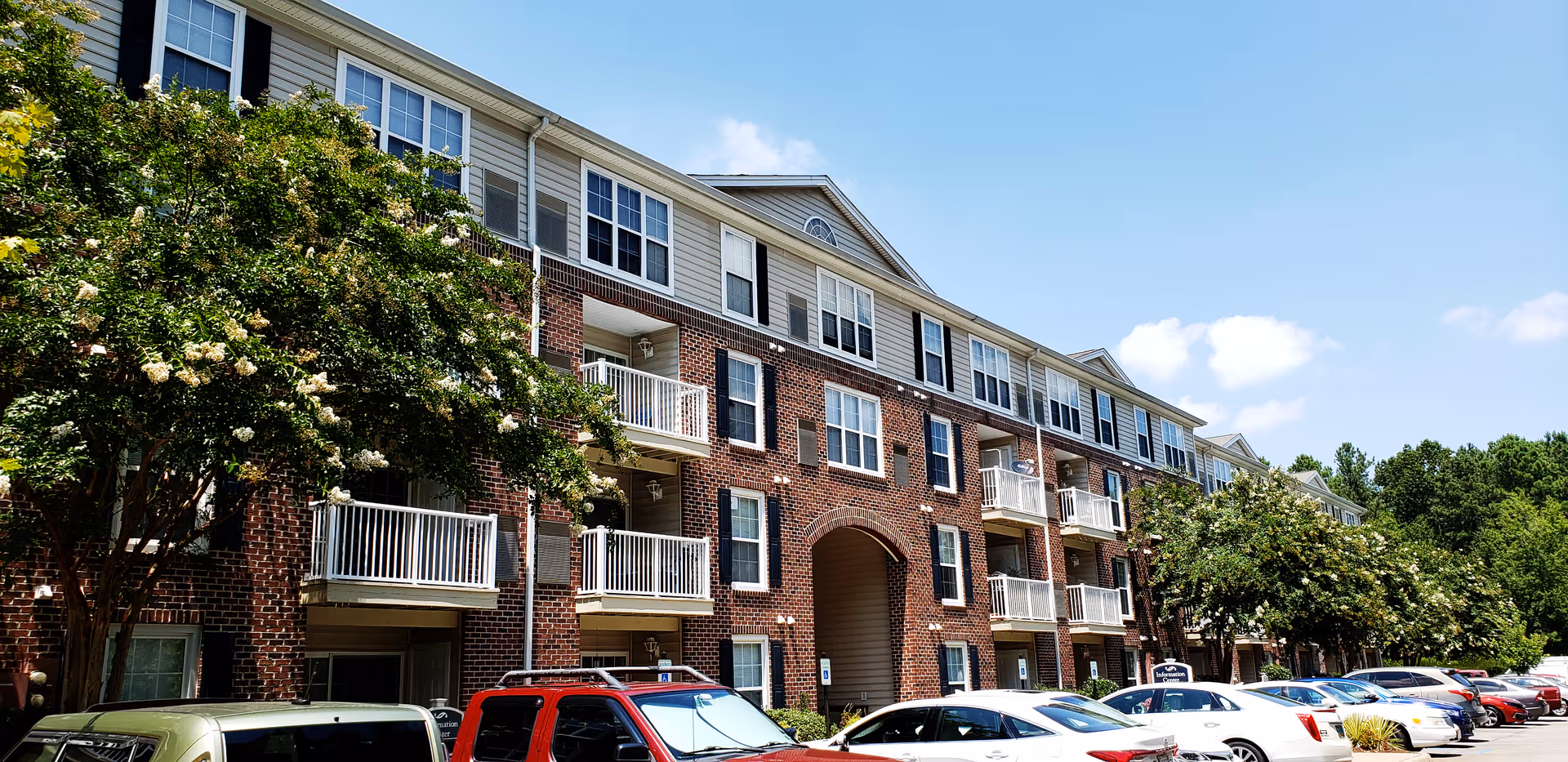 Three-story brick and siding apartment building with balconies, trees, and a row of parked cars under a clear blue sky.