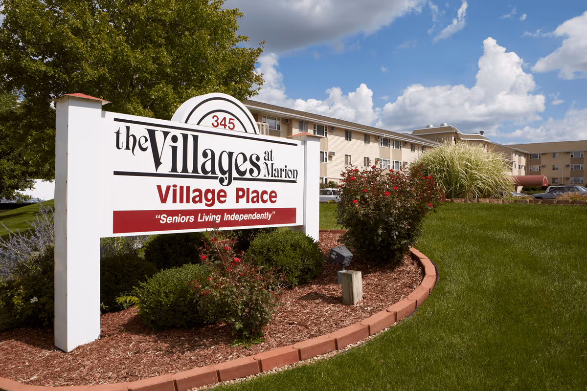 Entrance sign for Village Place in front of a multi-story senior living building with landscaping and a blue sky.