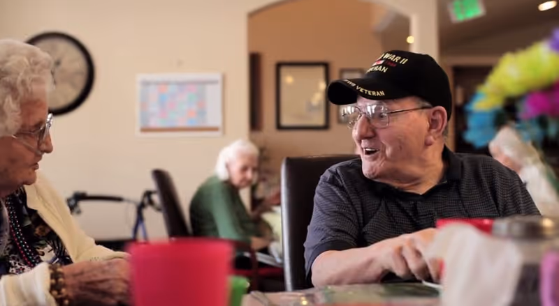 Several elderly residents sit and converse around a table in a communal dining area of a senior living facility.