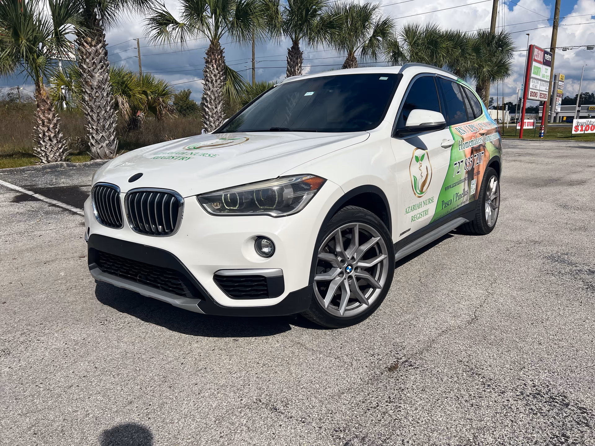 A white SUV branded with Azariah Nurse Registry parked in a lot, palm trees in background.