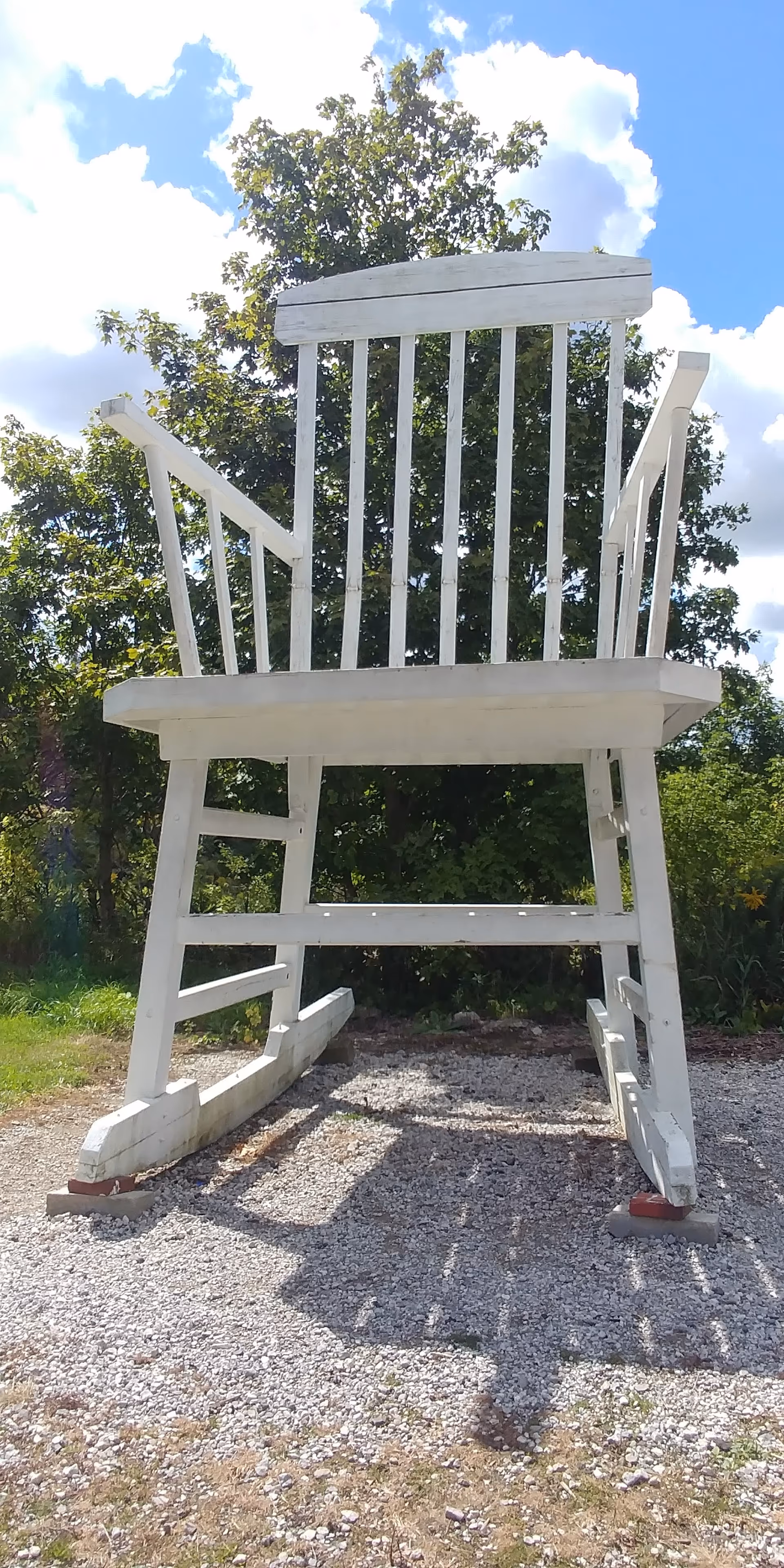 A giant white wooden rocking chair sculpture outdoors on gravel with trees and blue sky.