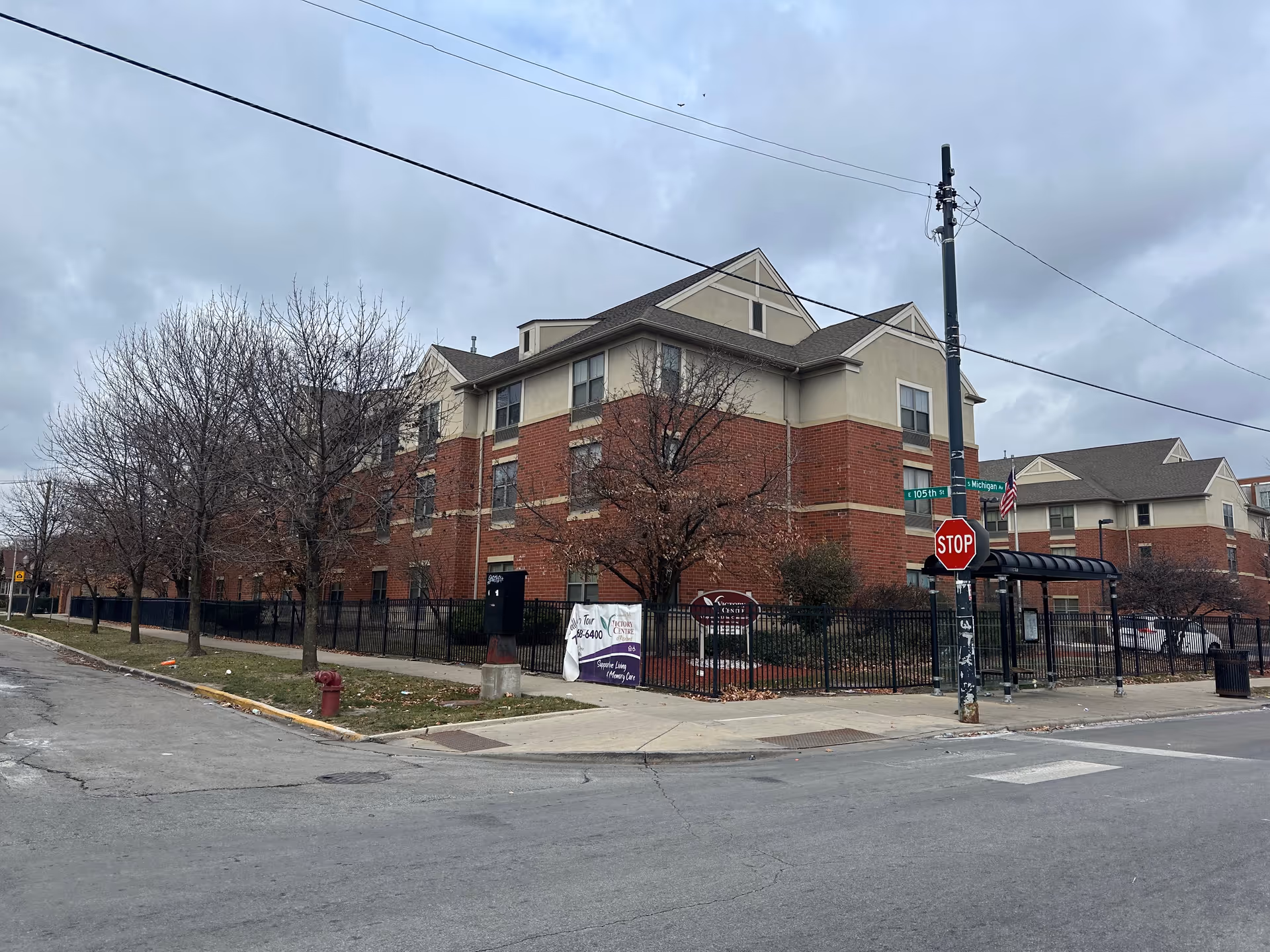 Brick three-story senior living building on a street corner with a fenced front, stop sign, and overcast sky.