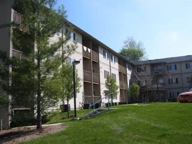 Exterior view of a multi-story residential building with balconies, surrounded by green grass and trees under a clear blue sky.