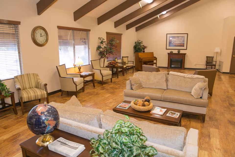 A bright common living area with sofas, armchairs, coffee tables, a piano and a faux fireplace under exposed ceiling beams.
