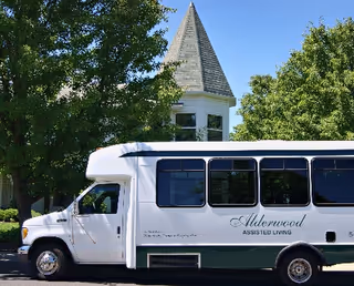 A white Alderwood Assisted Living shuttle bus parked in front of a stone building with trees.