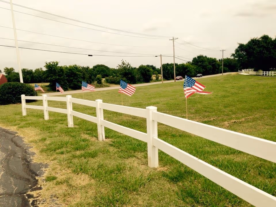 Green field with a white fence and multiple American flags along a rural road.