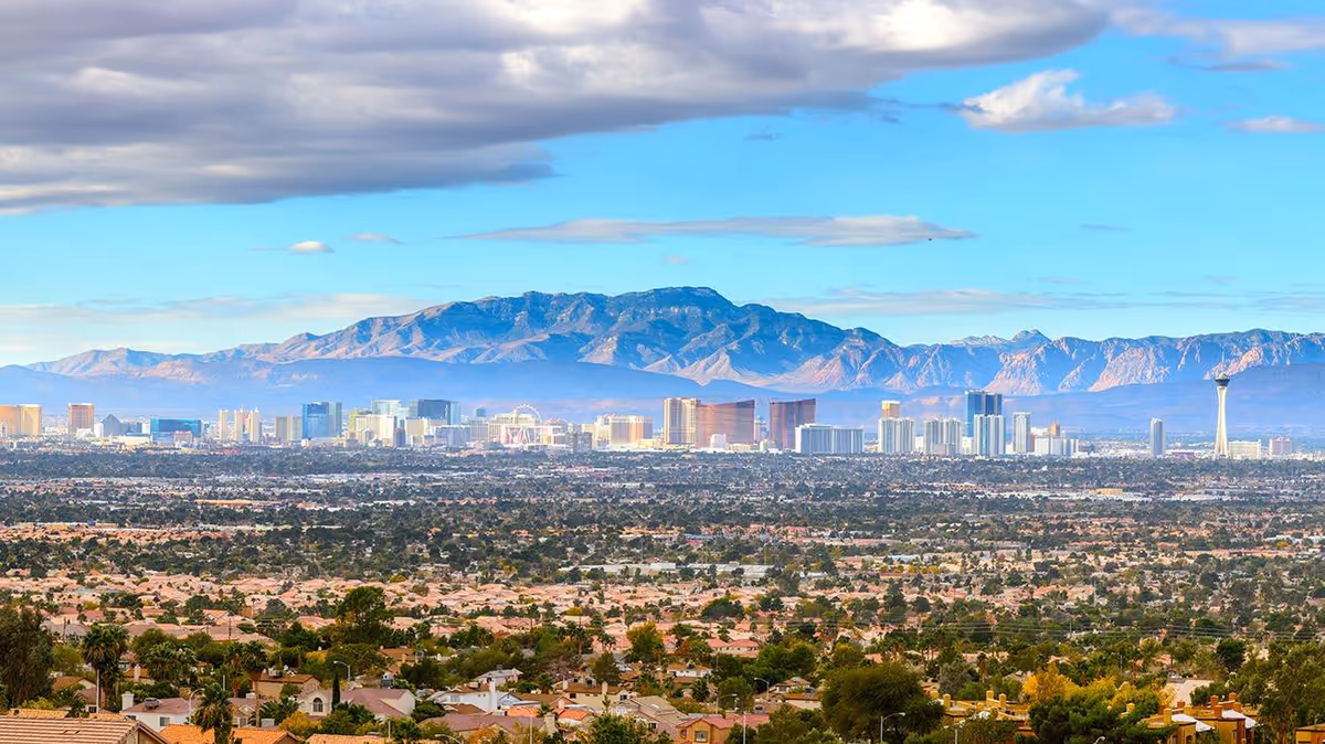 Panoramic view of the Las Vegas skyline with surrounding neighborhoods and mountains under a partly cloudy sky.