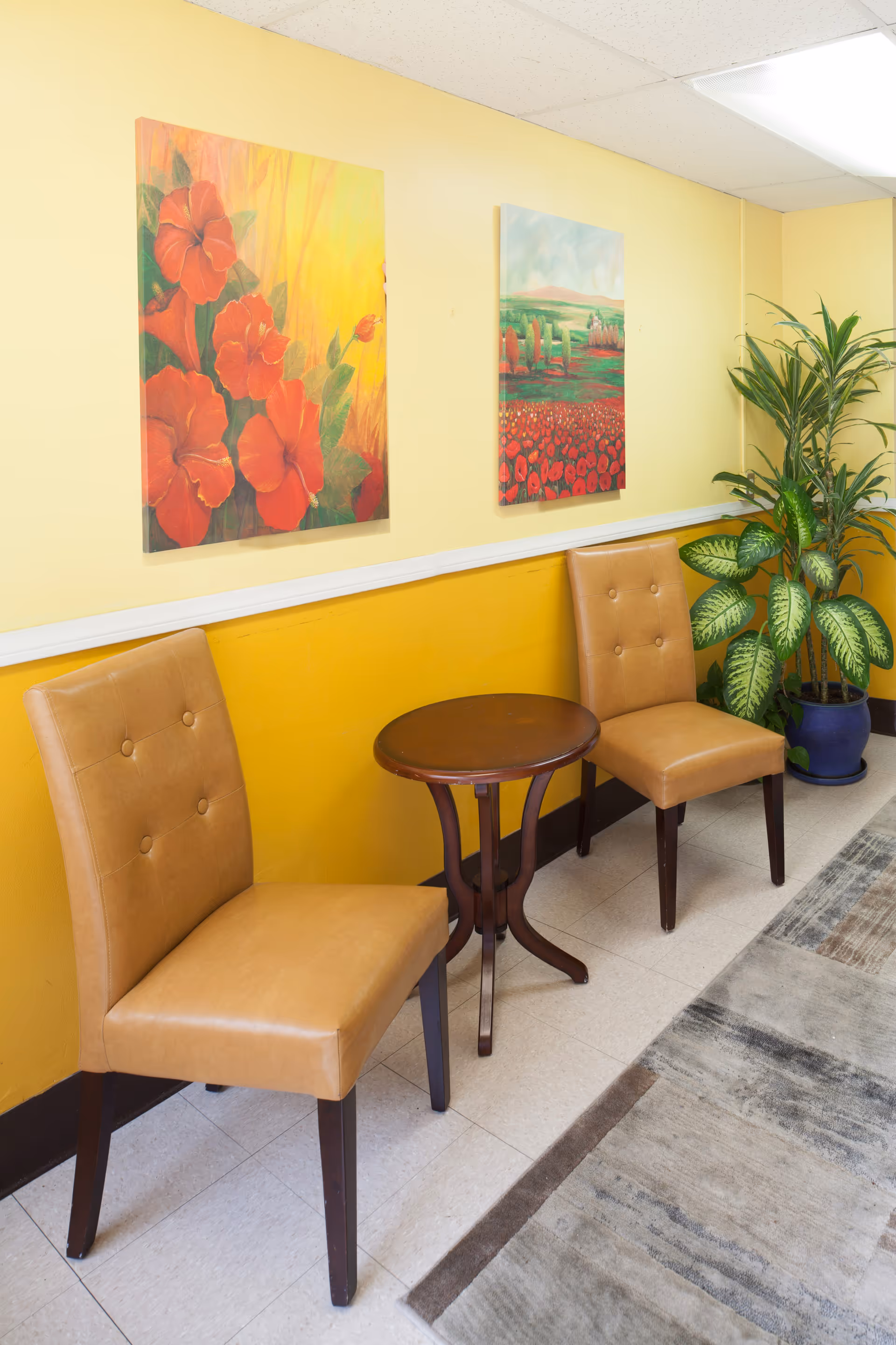 Yellow-walled lobby seating area with two tan chairs, a round table, plants, and paintings.
