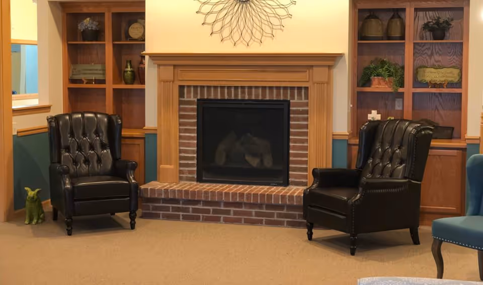 A cozy sitting area with two black leather tufted armchairs facing a brick fireplace with a wooden mantel. On either side of the fireplace are built-in wooden shelves displaying decorative items and plants. The walls are painted in a warm beige and teal color combination.