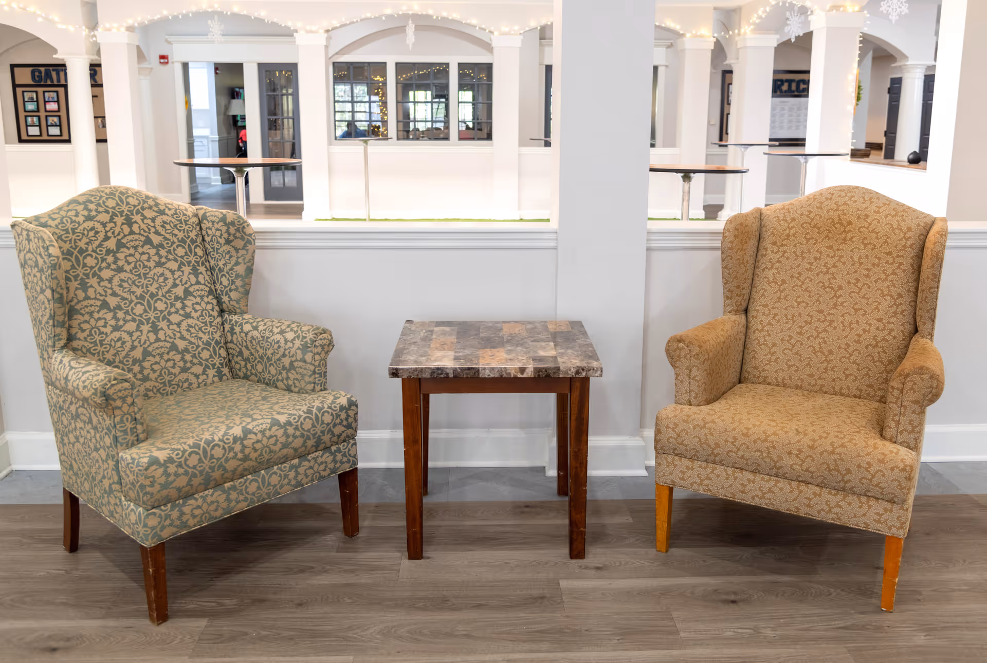 Two upholstered armchairs with patterned fabric, one greenish and one beige, positioned on either side of a small square wooden table with a tiled top, set against a white half-wall with windows and arches in the background, decorated with string lights.