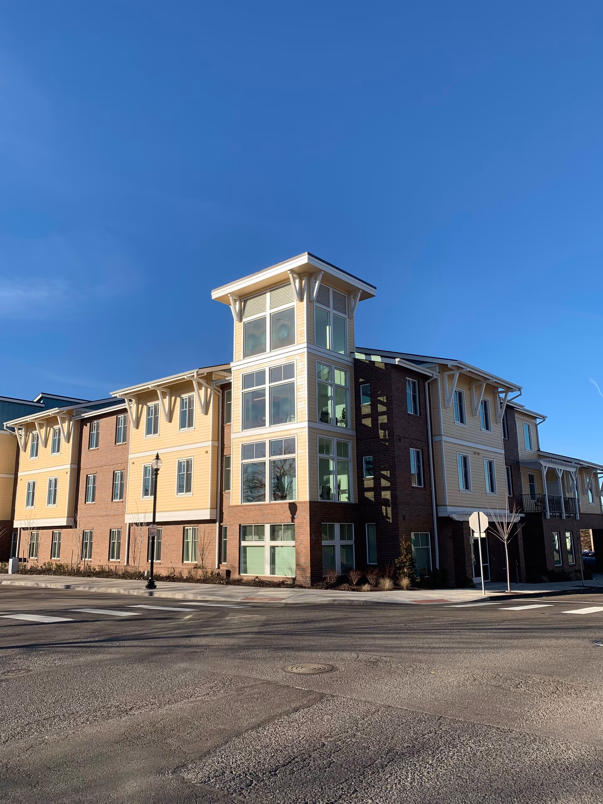 Three-story brick and yellow-clad senior living building with a glassed corner tower at a street intersection under a clear blue sky.