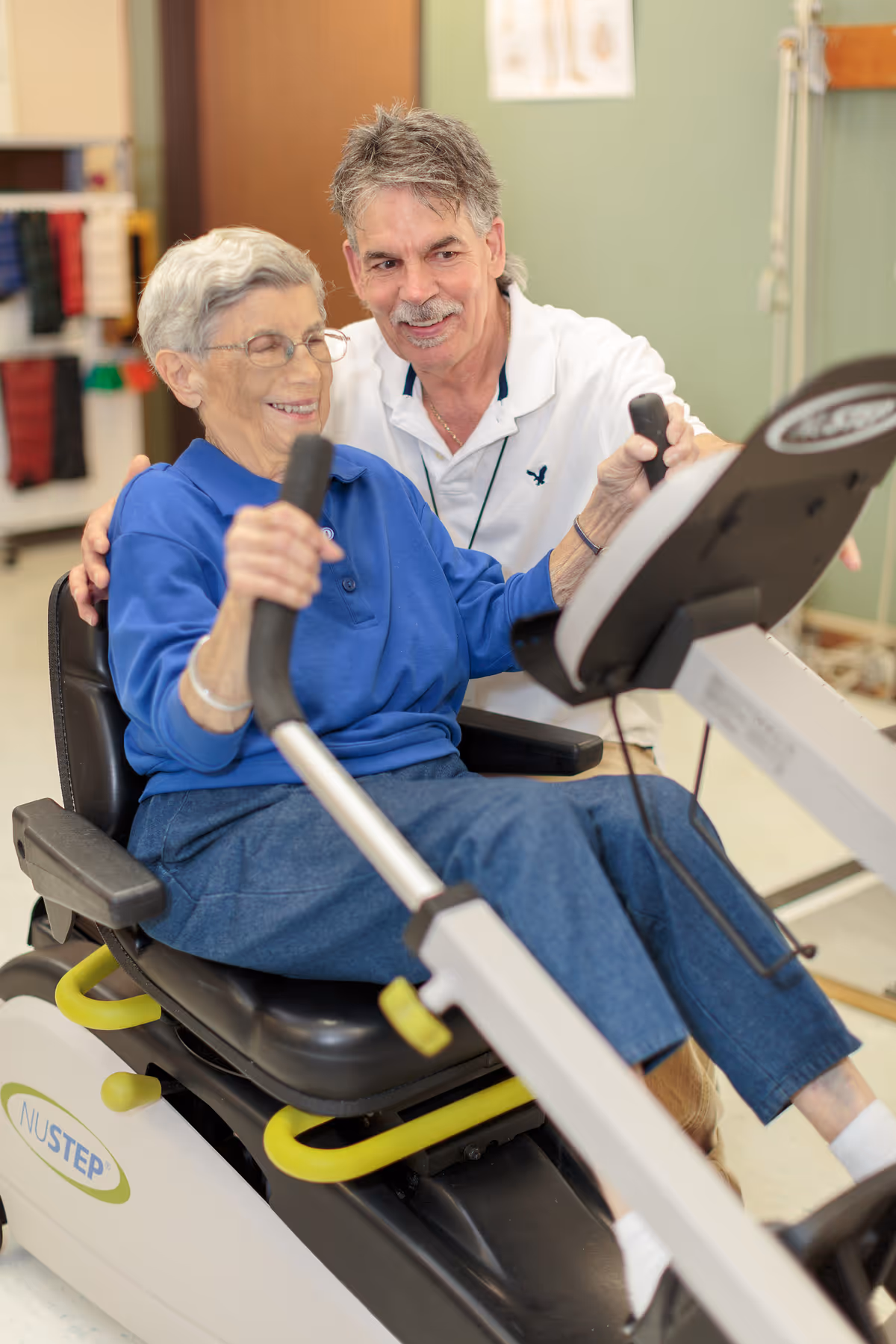 An elderly woman in a blue sweater and glasses is using a NuStep exercise machine while a man in a white shirt supports and encourages her. They are both smiling in a bright indoor setting.