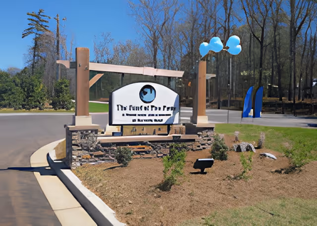 Entrance sign for The Pearl at Five Forks facility, mounted on a stone base with wooden posts, surrounded by a landscaped area with small shrubs and a paved road curving around it. Trees and a clear blue sky are visible in the background.