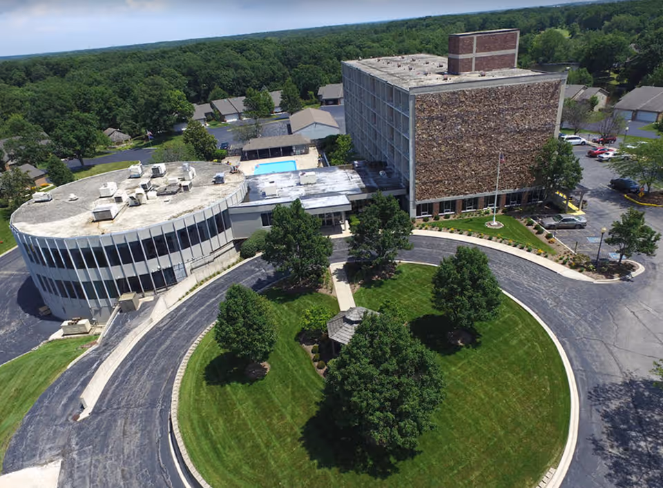 Aerial view of a senior living facility named Village Woods featuring a circular driveway with green lawns and trees in the center, a multi-story building with a stone facade, an adjacent curved building with large windows, and a swimming pool in the background surrounded by trees and residential houses.