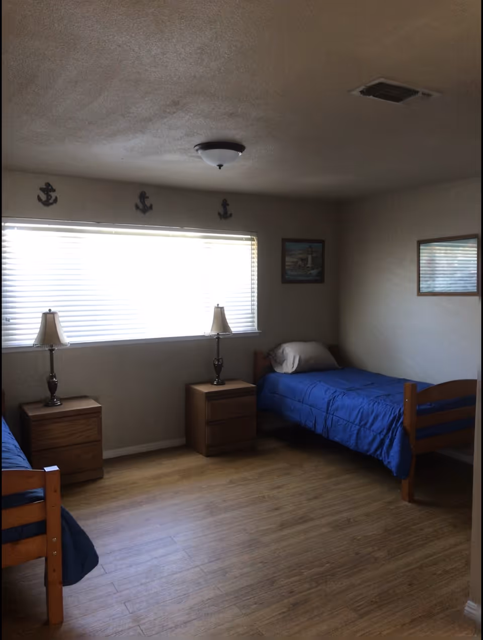 Simple shared bedroom with two single beds with blue bedding, wooden nightstands and lamps beneath a wide window.