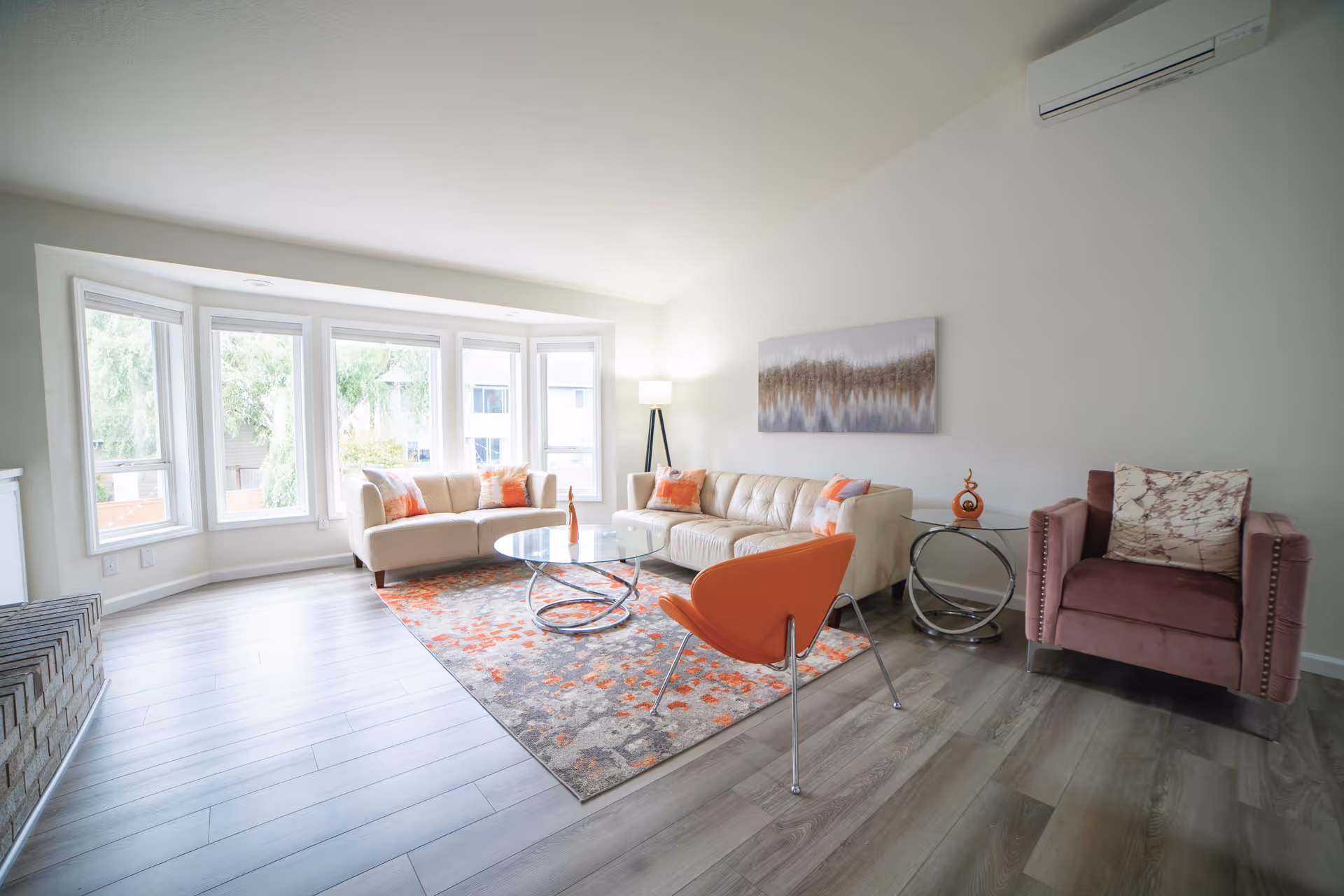 Bright living room with curved white sofa, orange chair, glass coffee table, and bay windows.