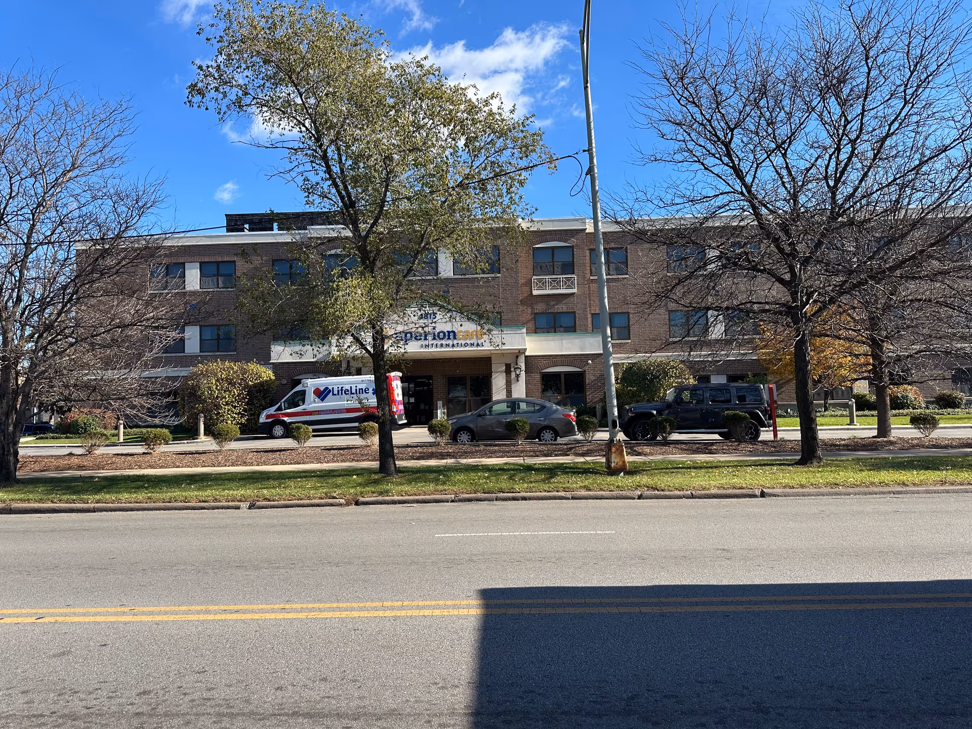 Three-story brick building entrance of Aperion Care International viewed from across the street with parked vehicles and trees in front.