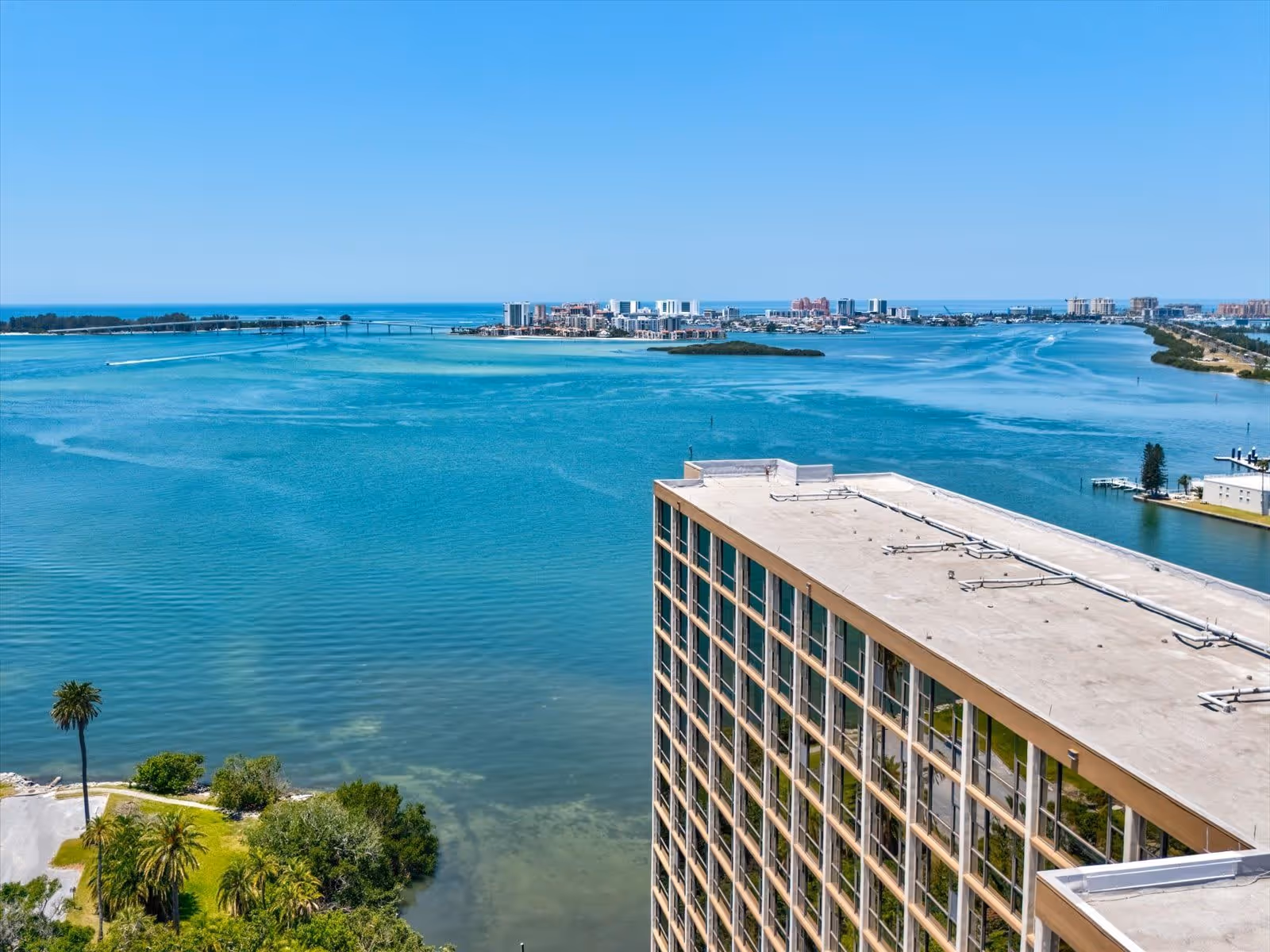 Aerial view of a large building next to a body of water with a clear blue sky. The water extends into the distance with a bridge and cityscape visible on the horizon. Palm trees and greenery are visible near the water's edge.