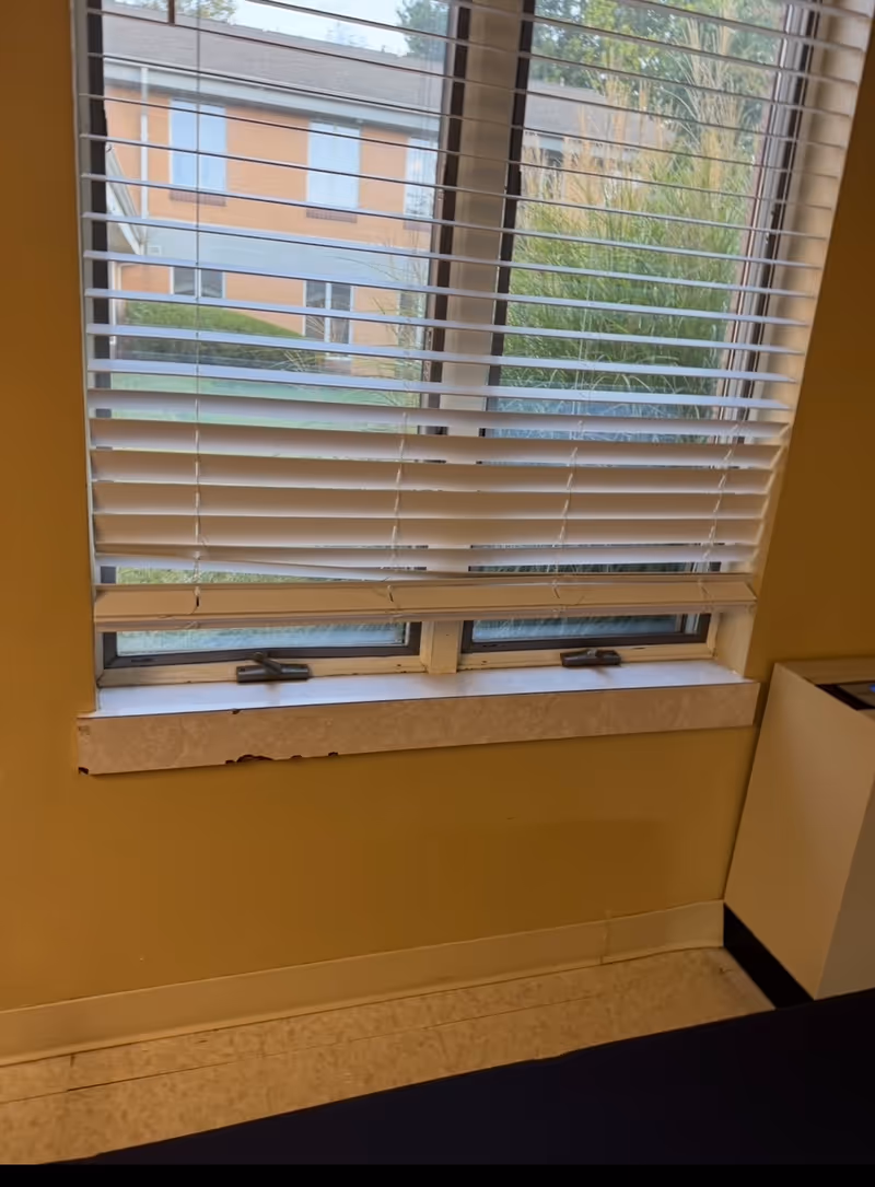 Indoor room with window and horizontal blinds, revealing an exterior brick building.