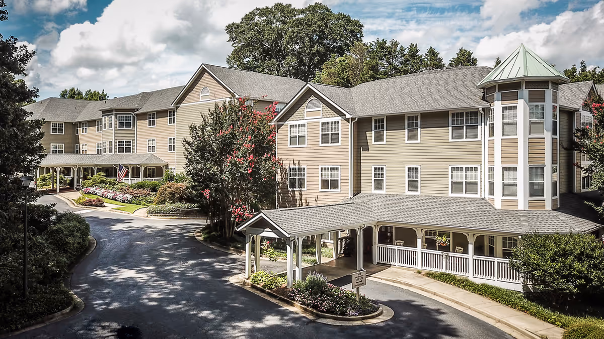 Exterior view of a large, multi-story retirement community building with beige siding and white trim. The building features a covered entrance with a driveway, landscaped gardens with flowering plants, and mature trees under a partly cloudy sky.