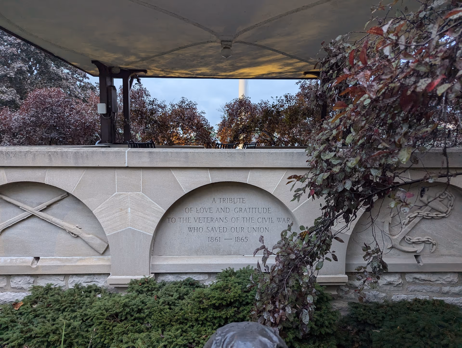 Stone exterior wall with a memorial plaque under a shaded canopy, surrounded by shrubs.